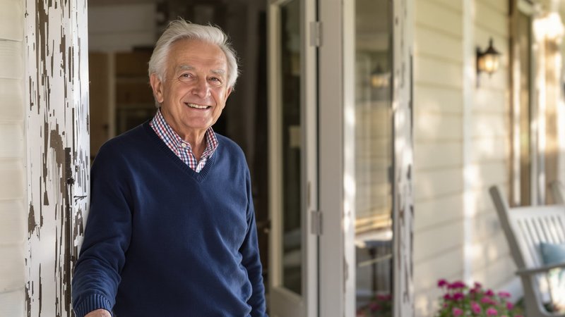 An older man on the front porch of his single-family home in soft afternoon light, with a gentle thoughtful smile.