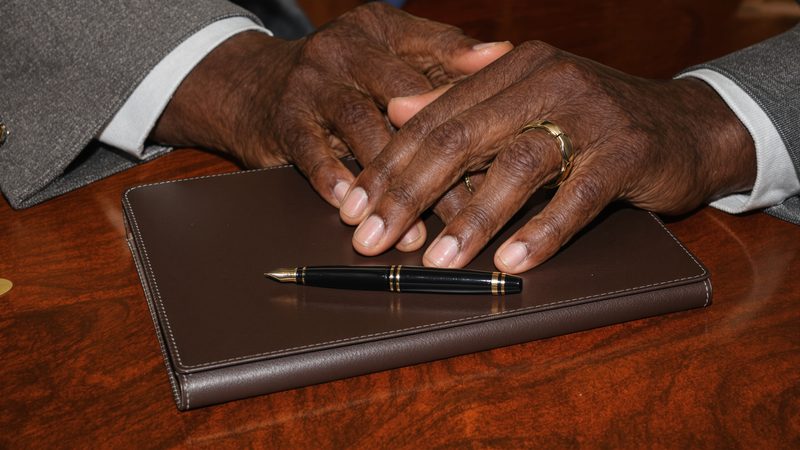 An older man's hands with a pen, about to sign a stack of legal documents on a desk — power of attorney and healthcare directives.