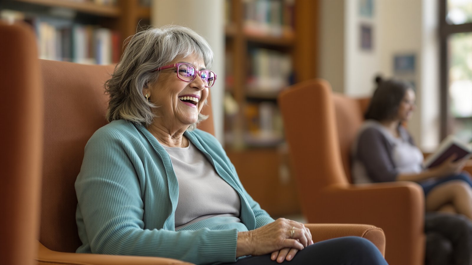 An older white woman with silver hair laughing mid-conversation in a community library reading nook.